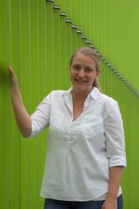 Lisa-Marie steht lächelnd vor den Glasrohren der Algenproduktion.